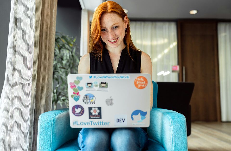 Smiling woman with red hair sitting in a teal armchair, using a laptop.