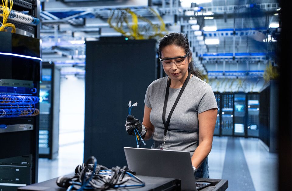 Woman in safety glasses holding network cables while working on a laptop in a data center.