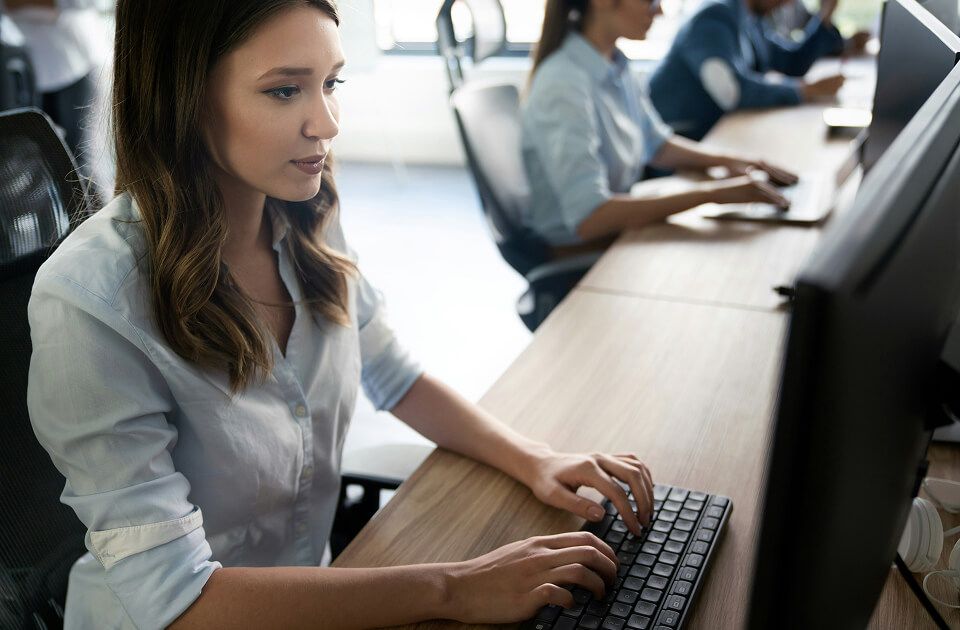 Woman typing on a keyboard while focused on a computer monitor, with coworkers working at desks in the background.