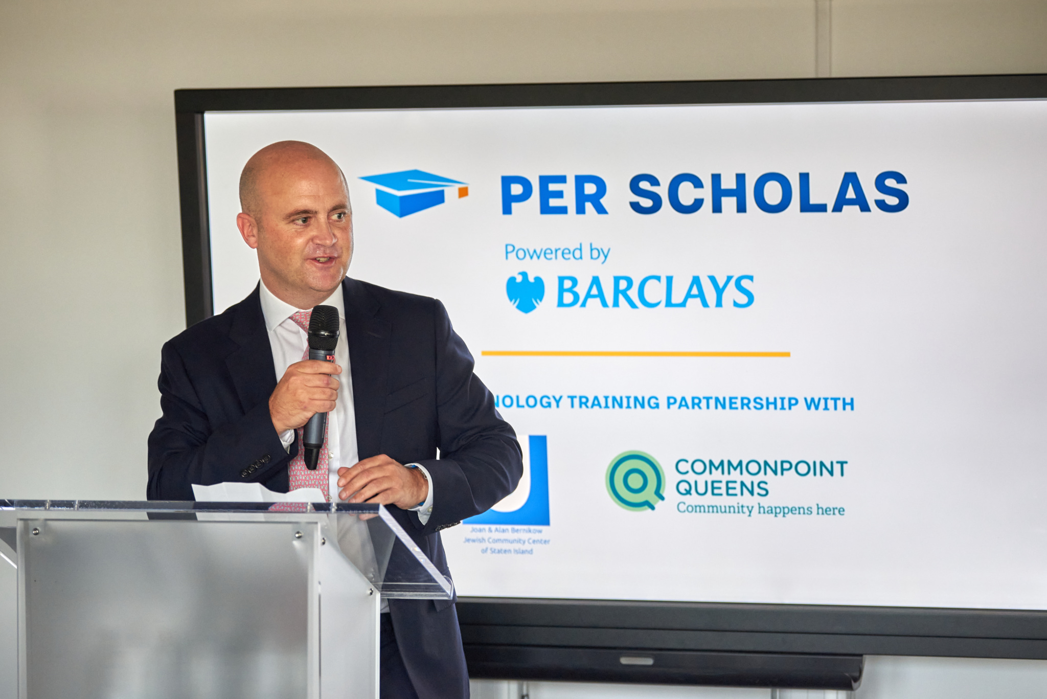 Man speaking at a podium with a microphone in front of a screen that displays "Per Scholas" and "Powered by Barclays" and partner logos.