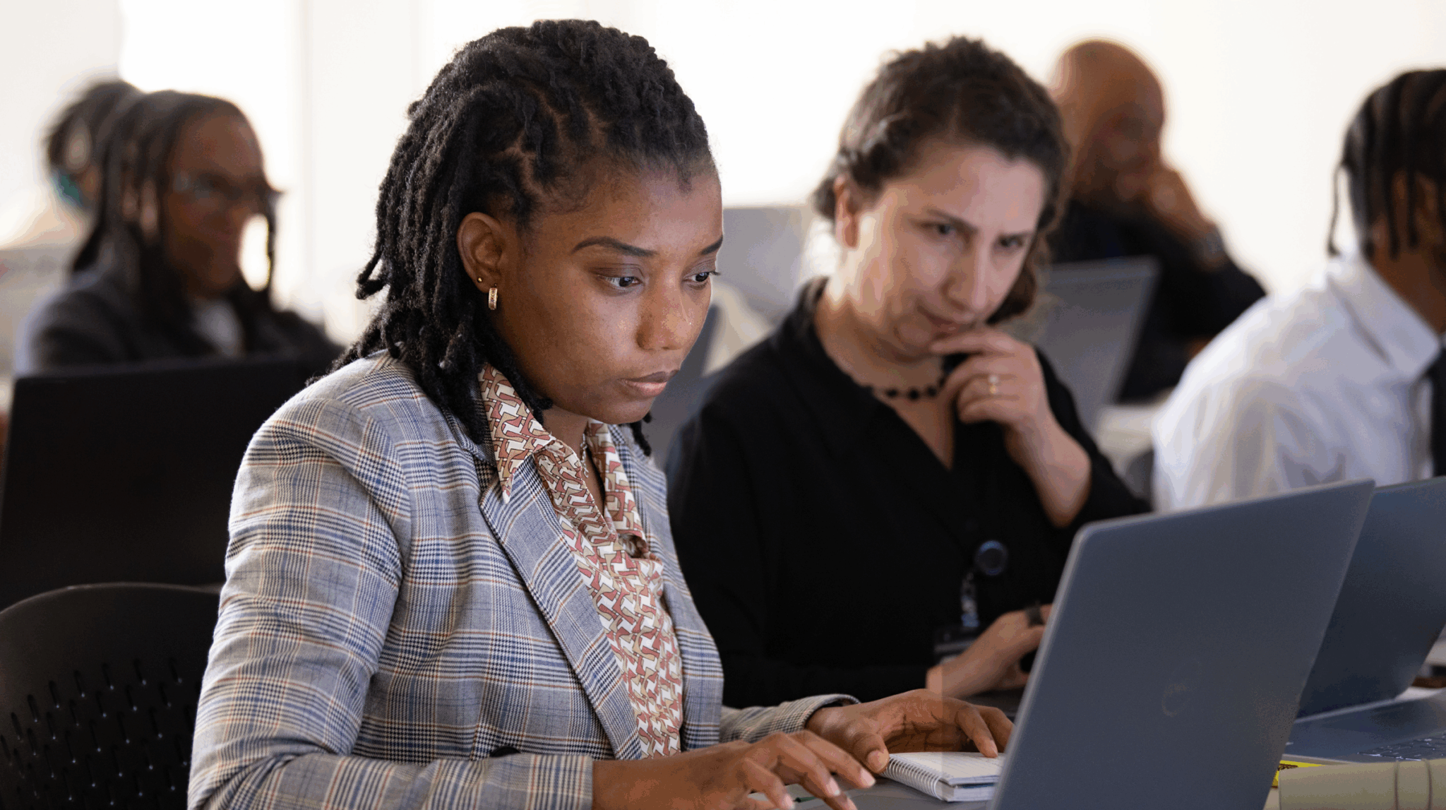 Two adult learners focused on laptops in a classroom, one typing while another looks on thoughtfully.