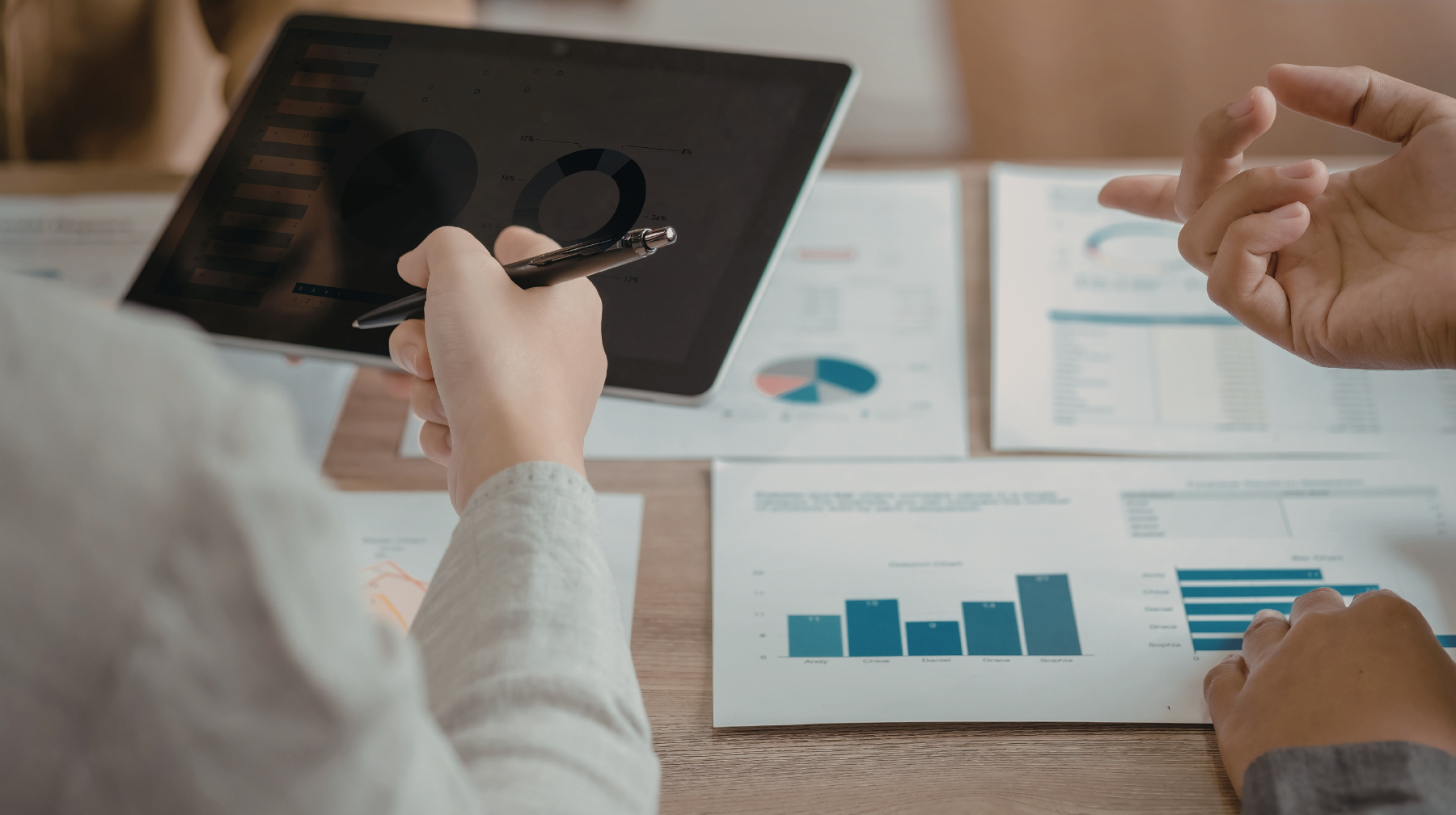 Two people reviewing charts and graphs on a tablet and printed reports at a table, pointing and discussing data.