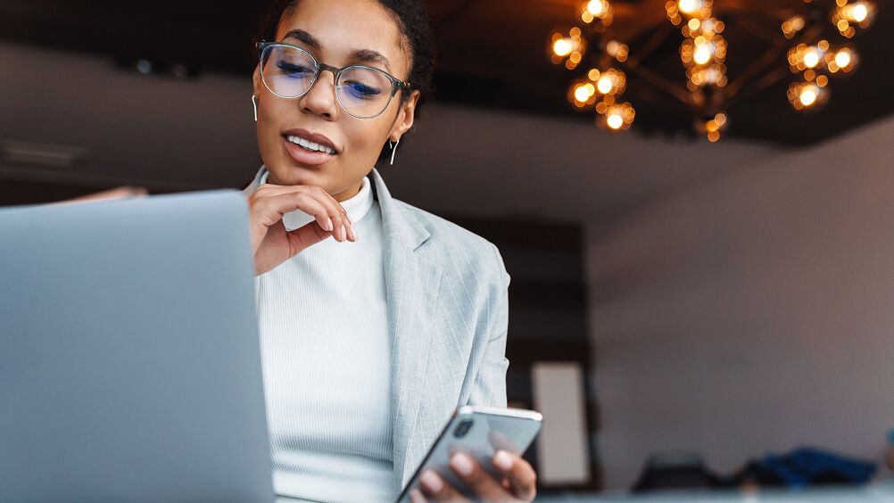 Smiling professional woman in glasses uses a laptop and checks her smartphone in a modern office, conveying focused, confident productivity.