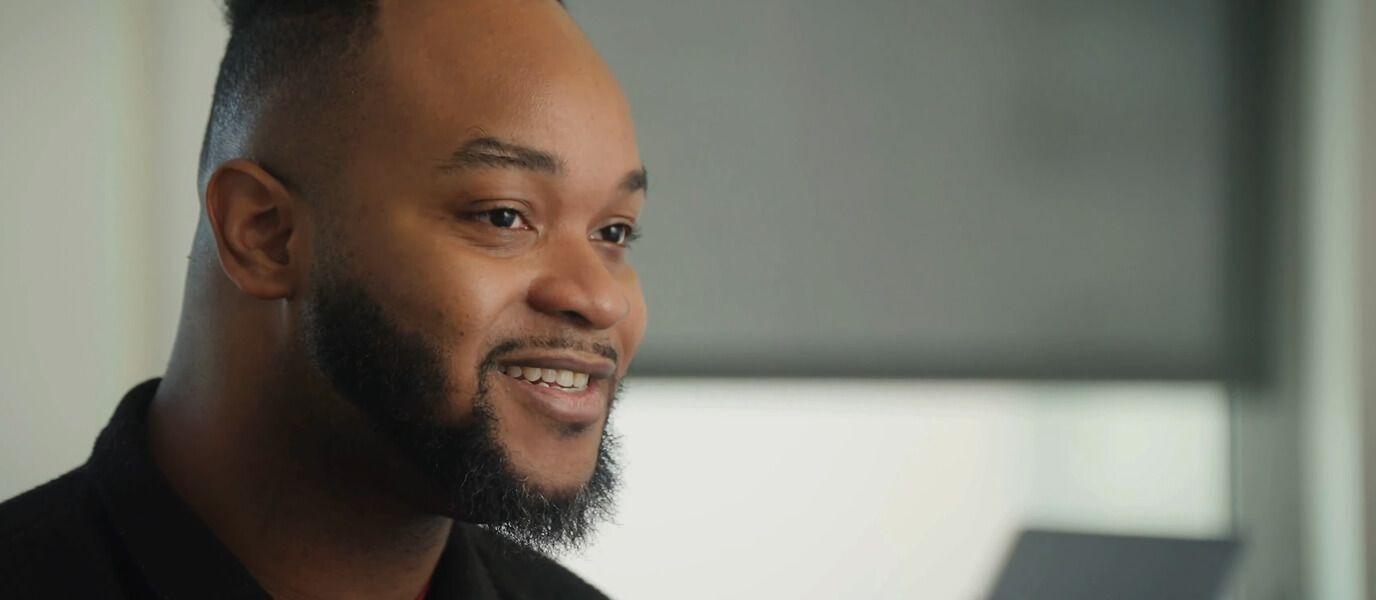 Smiling Black man with a beard sits in a modern office, looking toward a laptop; warm natural light creates a friendly, professional mood.
