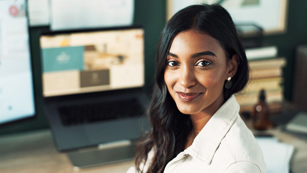 Smiling woman with long dark hair in a white shirt sits in an office beside computer screens, conveying warmth, confidence, and professionalism.