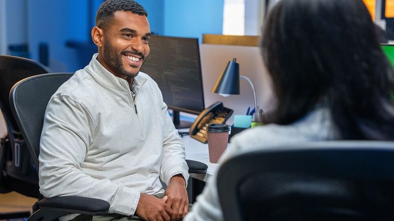 Smiling Black man in a modern office speaks with a colleague in the foreground, creating a friendly, professional atmosphere.
