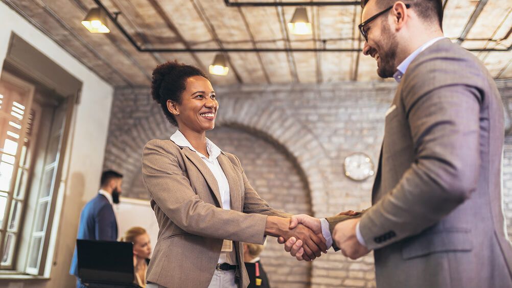 Businesswoman and businessman shake hands and smile in a bright brick office, conveying a warm, friendly professional meeting at work.