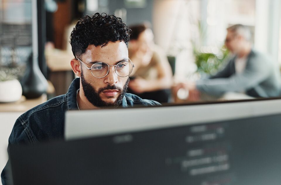 Focused young man intently works on a laptop, with blurred coworkers behind him.