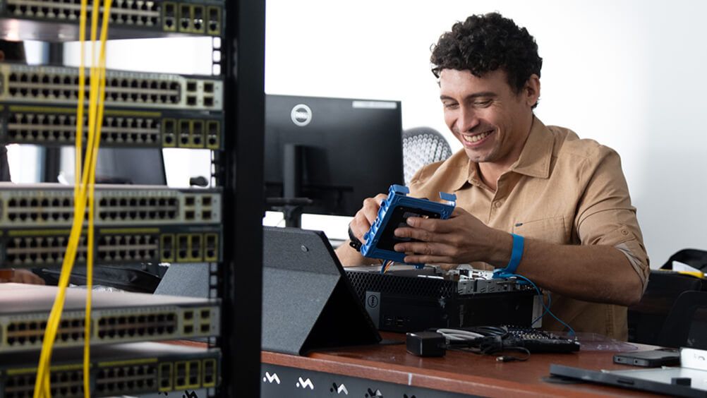 Technician smiling while repairing network hardware with a blue tool at a workbench beside server racks and yellow cables in an office.
