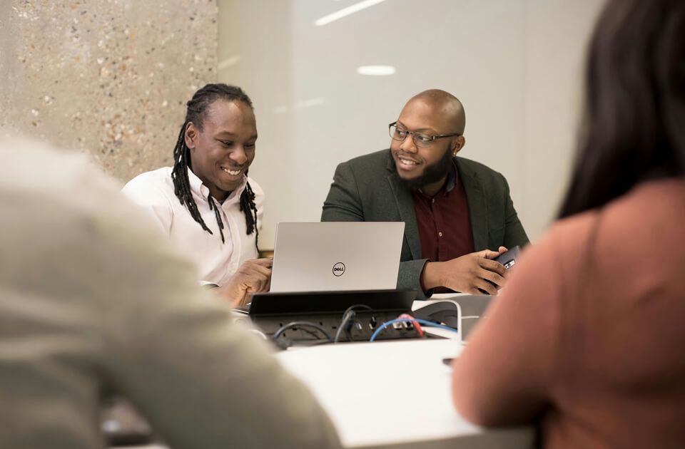 Two men smiling while looking at a laptop together, with two colleagues seated in the foreground at a conference table.