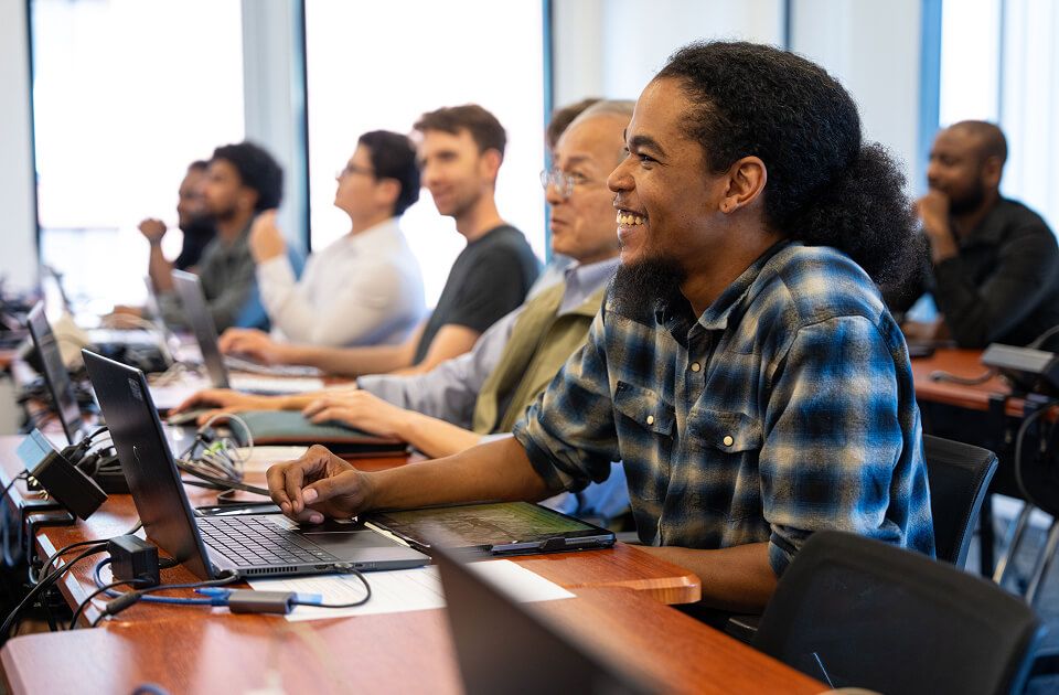 Man with curly hair smiling while working on a laptop, seated in a row with several other attentive learners at desks.