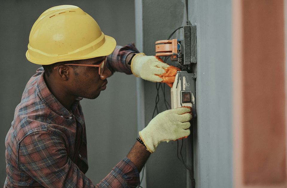 Man wearing a yellow hard hat, safety glasses, and gloves closely inspecting and wiring an electrical outlet on a concrete wall.