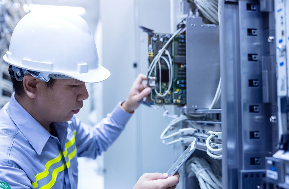 Technician wearing a white hard hat inspecting a circuit board panel inside a server cabinet while holding a tablet.