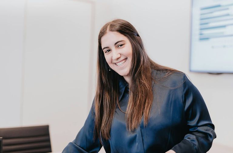Smiling woman with long brown hair in a navy blouse leans toward the camera in a bright office, conveying warmth and confidence.