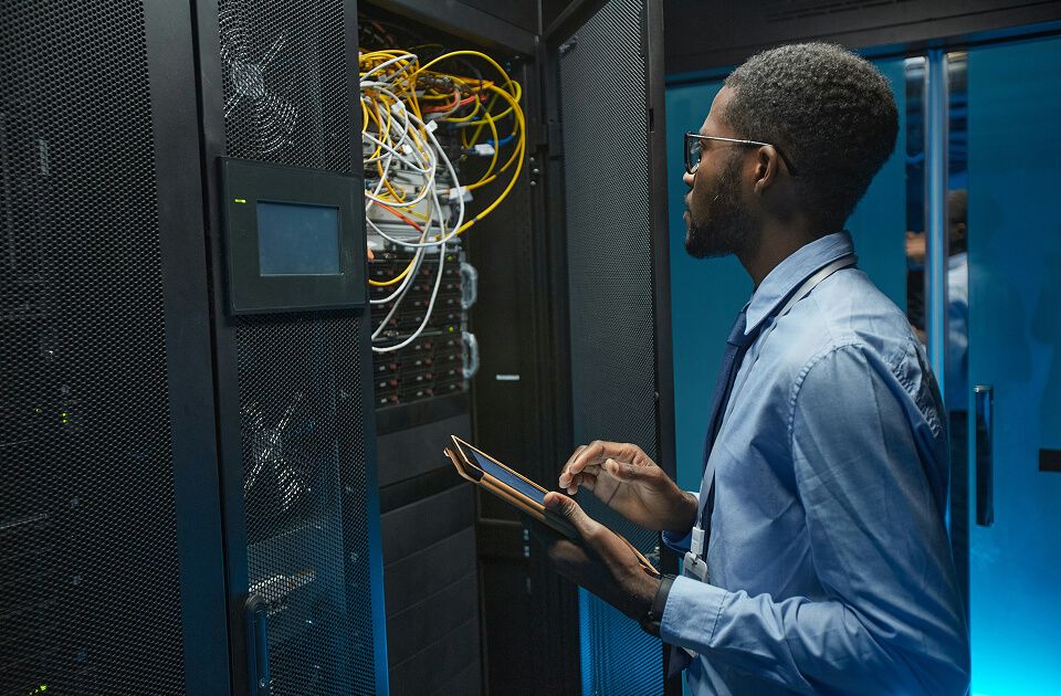 Man wearing glasses and a dress shirt holding a tablet while inspecting an open server rack with yellow and white cables in a blue-lit data center.