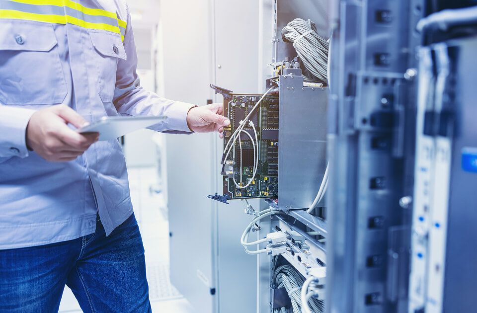 A male technician holding a tablet while inspecting and connecting cables on a circuit board module within a data center server rack.