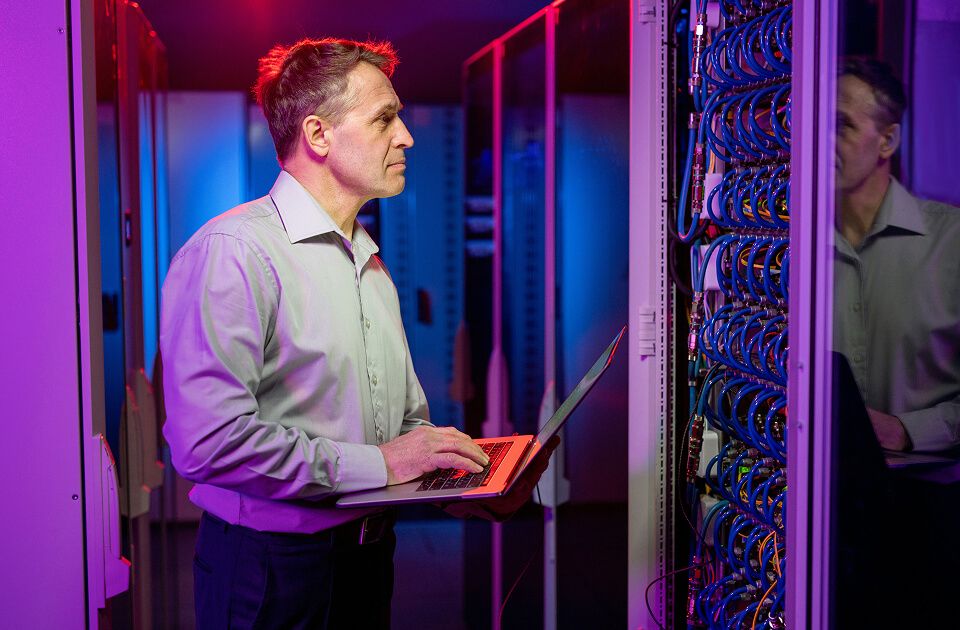 Man holding a laptop while inspecting server racks with blue cabling in a data center, illuminated by purple and blue lighting.
