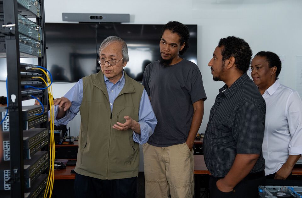 Male instructor gesturing while explaining server rack wiring to three attentive learners in a classroom setting.