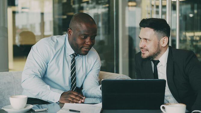 Two men in professional attire in conversation over a laptop during a meeting in an office.