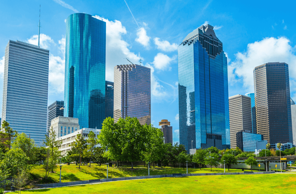 Downtown Houston skyline rising above a green park under a bright blue sky.