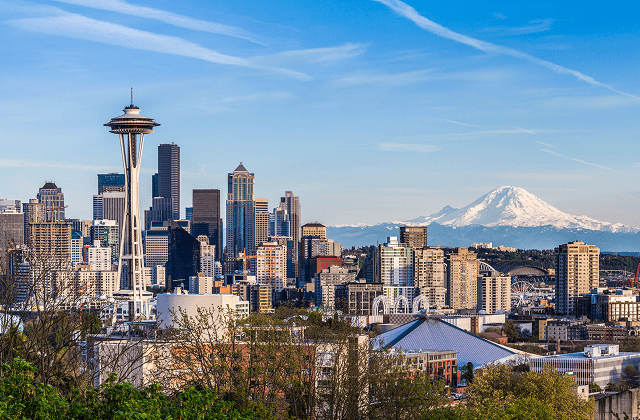 Seattle skyline with the space needle and downtown skyscrapers, with a snow-capped mountain visible in the distance under a clear blue sky.