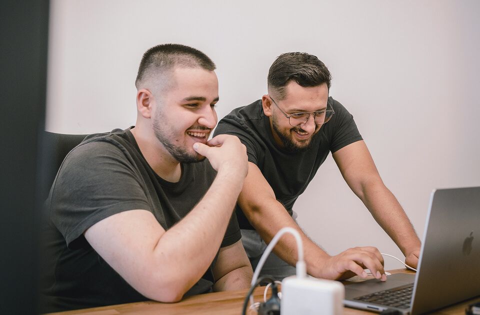 Two men in dark t-shirts smiling and laughing while looking at a laptop screen together.