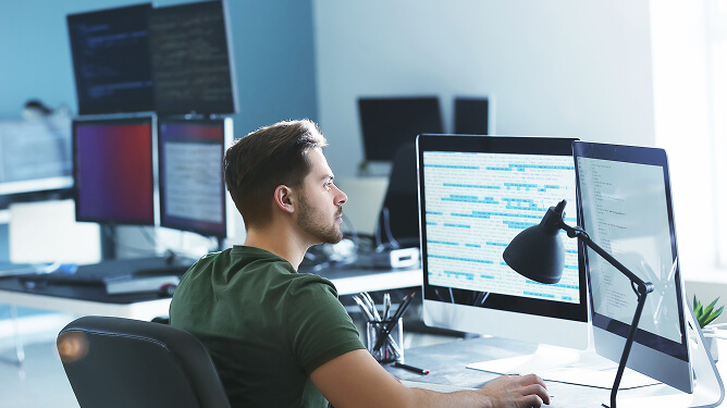 Man at a workstation focused on code displayed across multiple monitors in an office setting.