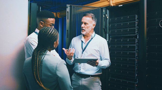 Instructor holding a tablet while speaking to two employees beside server racks.