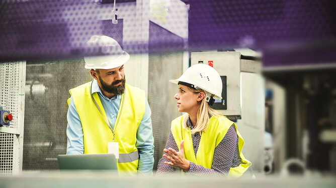Two safety-helmeted mentors talk over equipment on a factory floor.