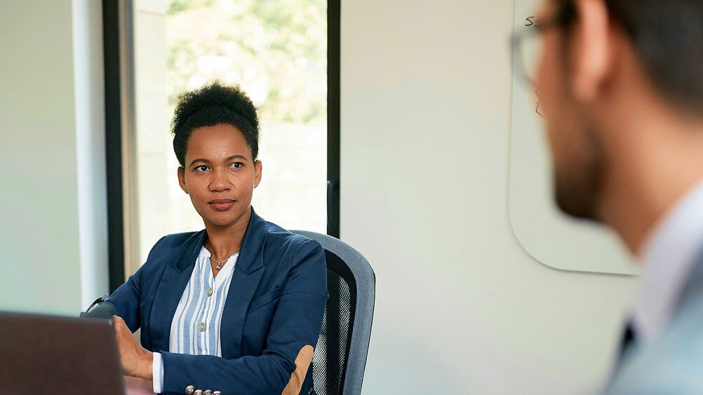 Professional Black woman in a blue blazer sits at a laptop in an office, listening attentively to a blurred male colleague during a discussion.