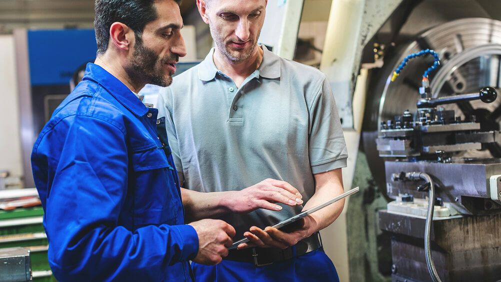 Two engineers in a factory review a tablet beside industrial machinery, collaborating closely; one wears blue workwear, the other gray.