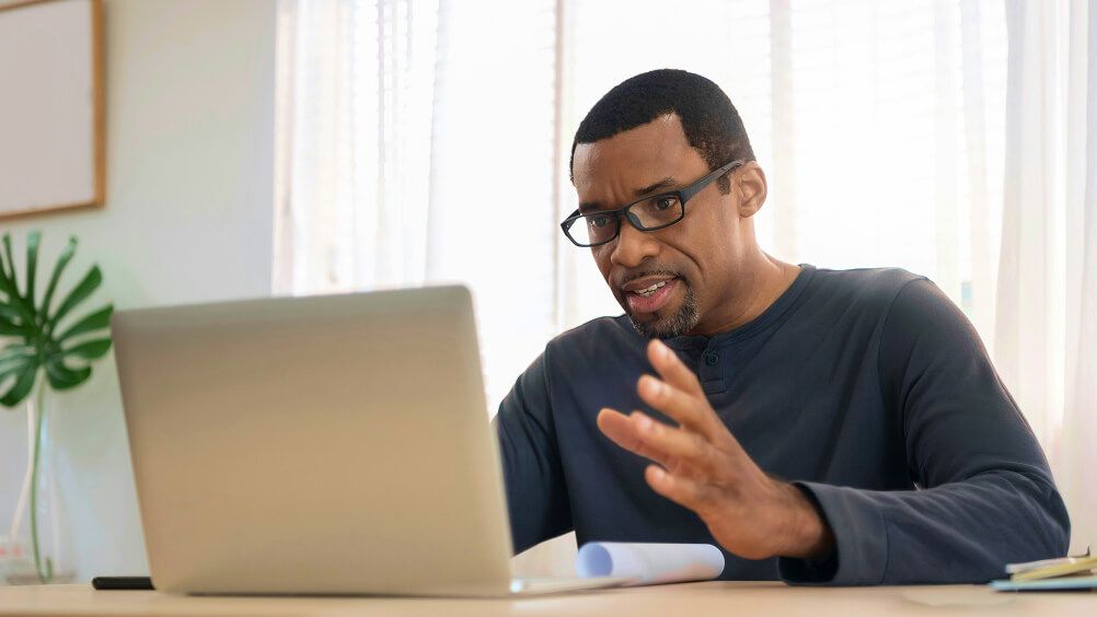 A man in glasses gestures beside a laptop at home, appearing engaged and thoughtful during a video call or focused online work session.