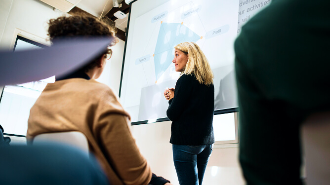 Woman standing at a projector screen teaching to seated adult learners.