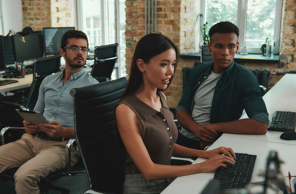 Woman typing on a keyboard in the foreground while two men sit behind her at desks with computer monitors in a brick-walled office.