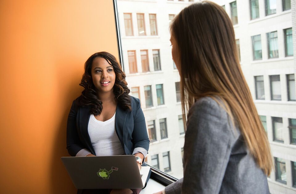 Two female professionals working on laptops by a window in an office building. 