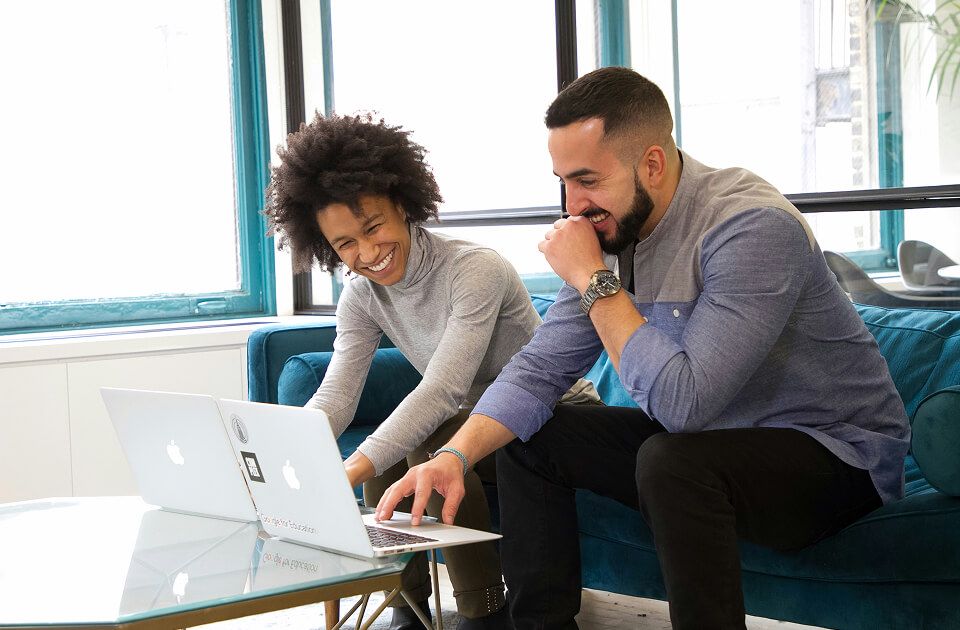 Man and woman laughing while looking at a laptop screen together, seated on a teal couch in an office lounge.