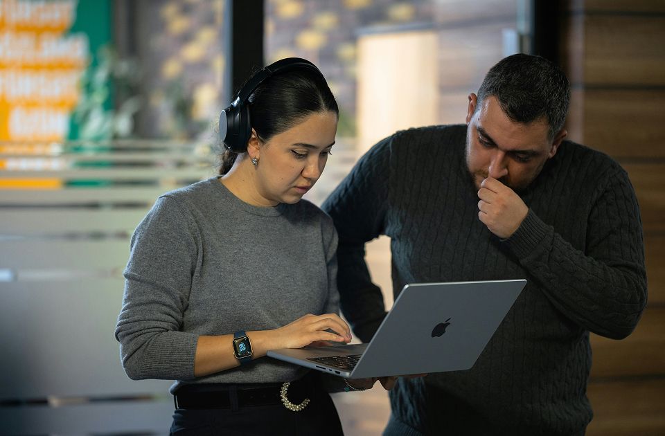 Woman wearing headphones typing on a MacBook while a man beside her looks on thoughtfully, standing in an office lobby.