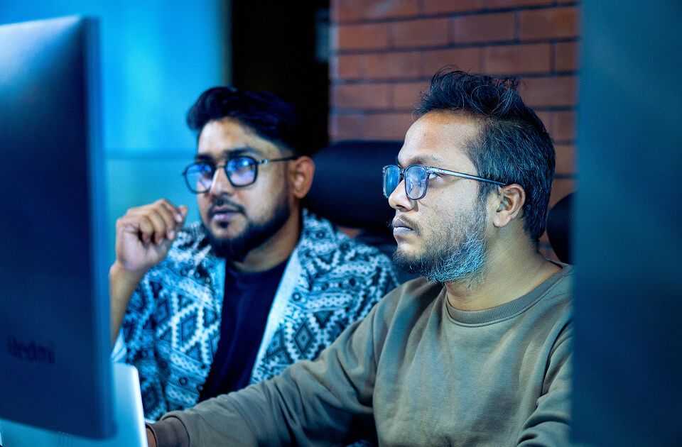 Two men with glasses focused intently on a computer monitor, one gesturing thoughtfully, in a dimly lit office with a brick wall background.