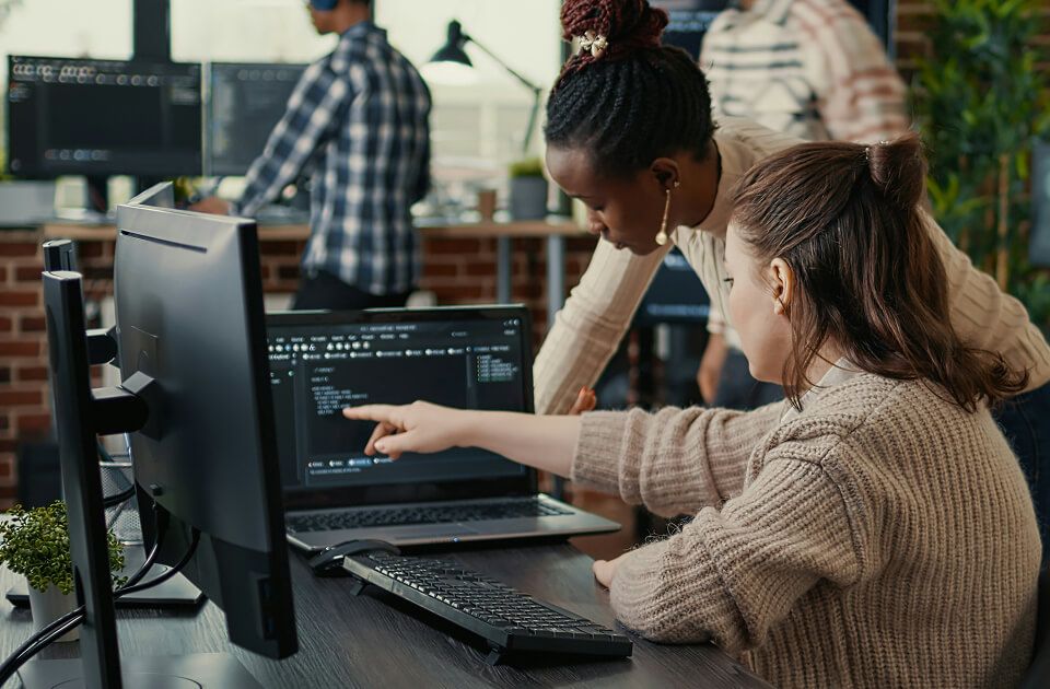 Two women reviewing code on a laptop and monitor, with one pointing at the screen, in an office with coworkers in the background.