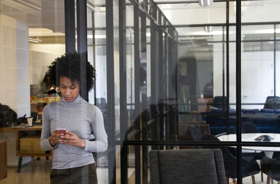A young professional standing in a modern office, looking at a smartphone near glass-walled meeting rooms.