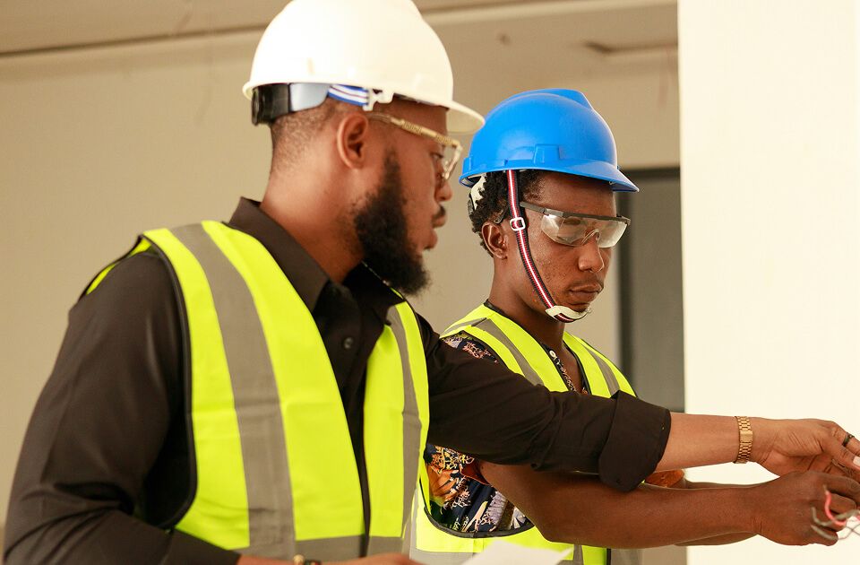 A senior professional in a white hard hat and safety glasses guides a younger professional in a blue hard hat and safety goggles, both wearing high-visibility yellow vests, as they inspect wiring.