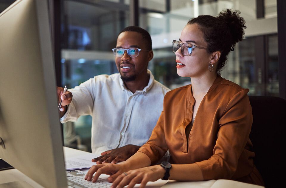 Man and woman wearing glasses looking at a computer monitor together in an office, with the man pointing at the screen.