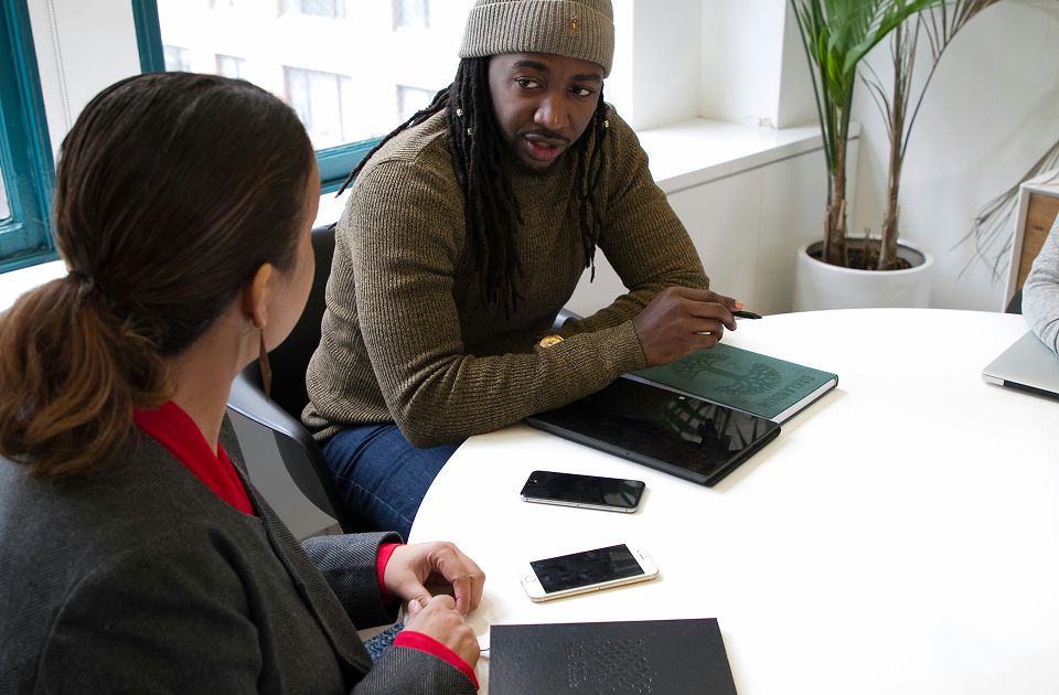 Man in a beanie and sweater speaking to a woman listening, with phones and a notebook on the table.